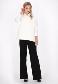 Maglione senza maniche color avorio con scollo a V, indossato sopra una camicia bianca con bottoni, abbinato a pantaloni a gamba larga neri a vita alta.