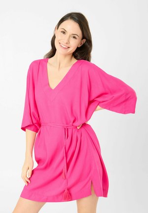 Femme aux longs cheveux foncés souriante, portant une robe ample rose vif avec un col en V et une ceinture à pompons, debout devant un fond blanc uni.