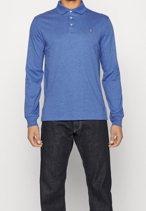 Modrá polokošile s dlouhým rukávem z měkké tkaniny, s límečkem na tři knoflíky a malým logem na prsou. Nošena s tmavými džínami.