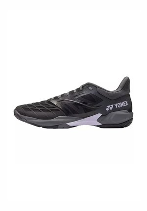 Zapato deportivo Yonex negro y gris con paneles de malla, suela texturizada y logo de la marca en el lateral, diseñado para el rendimiento deportivo.
