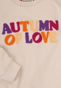 Getextureerde, meerkleurige letters die "HERFST VAN LIEFDE" spellen op een beige stof, met een opgevouwen mouw die zichtbaar is in de linkeronderhoek.