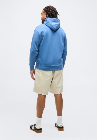 Άνδρας με dreadlocks που φοράει μπλε φούτερ με κουκούλα, μπεζ σορτς, λευκές κάλτσες και ασπρόμαυρα αθλητικά παπούτσια, στέκεται με την πλάτη προς τα εμπρός σε λευκό φόντο.