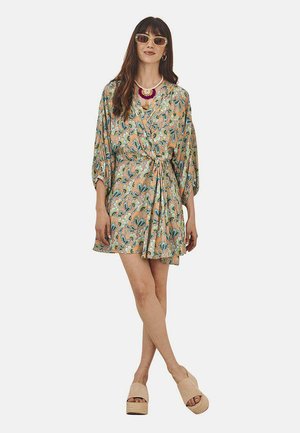 Vestido envolvente con estampado floral en beige con acentos en verde y azul; mangas largas y sueltas y cintura ceñida; combinado con sandalias de plataforma.