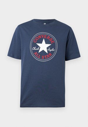 Navy plava majica kratkih rukava s bijelim logotipom zvijezde u sredini, okruženim crvenim tekstom "CONVERSE ALL STAR" i rukopisnim natpisom "Chuck Taylor".