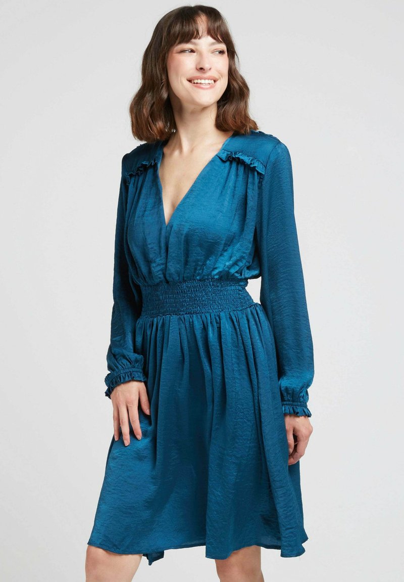 Robe teal avec un décolleté plongeant, des manches longues et une taille smockée. Présente des accents volantés sur les épaules et une jupe fluide. Texture soyeuse.