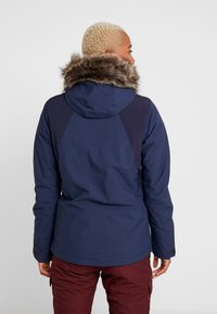 Giacca isolante blu navy con cappuccio foderato in pelliccia sintetica, caratterizzata da accenti scuri sulle spalle e da un design pratico e aerodinamico.