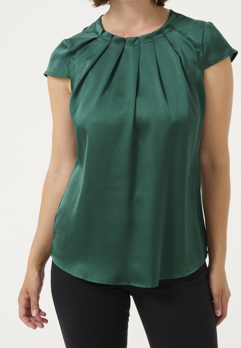 Femme portant un chemisier en satin vert foncé à manches courtes avec un col plissé, associé à un pantalon noir, sur un fond uni.