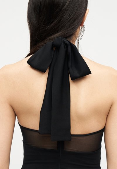 Femme aux longs cheveux noirs portant un haut dos nu noir noué en un nœud à l'arrière du cou, et une boucle d'oreille scintillante pendante à une oreille.