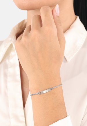 Bracciale d'argento con una placca centrale liscia e piatta e una catena intrecciata. Presenta una piccola foratura a forma di cuore da un lato.