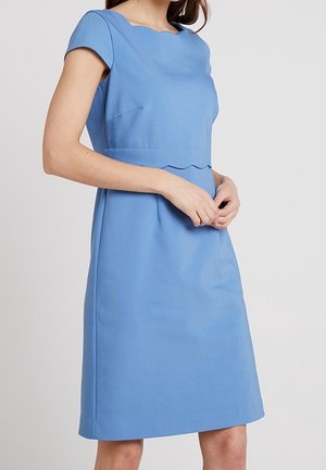 Mujer con un vestido azul hasta la rodilla, de manga corta, escote cuadrado y detalle en la cintura con bordes festoneados, de pie con las manos entrelazadas.