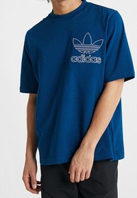 Mörkblå bomullströja med kort ärm, rund halsringning och en broderad vit Adidas-logga på vänster bröst.