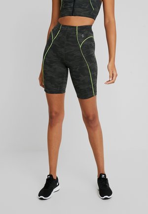 Femme portant un short de cyclisme camouflage noir avec des coutures vert néon et des chaussures de course Nike noires, debout sur un sol gris clair.