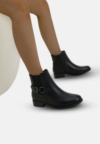 Piernas vestidas con botas de cuero negro hasta el tobillo con hebillas laterales y cremalleras, sentadas sobre una superficie curva blanca contra un fondo liso.