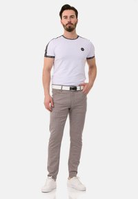 Weiße T-Shirt mit schwarz gestreiften Ärmeln, graue Nadelstreifenhose, weißer Gürtel und weiße Sneakers. Das Model steht mit einer Hand auf der Hüfte.