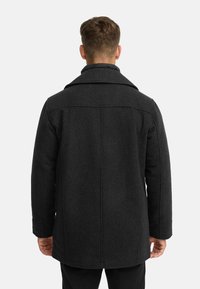 Svart ullfrakk med en strukturert design, bred krage og glatt tekstur. Legg merke til sømdetaljene og lengden som når midt på hoften.