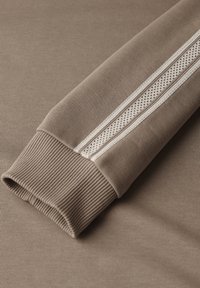 Tessuto di felpa marrone con polsini a costina presenta una striscia orizzontale bianca perforata lungo il braccio. Texture liscia, design minimal.
