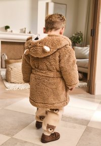 Kleinkind, das drinnen läuft und einen gemütlichen, braunen Bären-Haubenmantel sowie beige Hosen mit Bärenmotiven trägt.