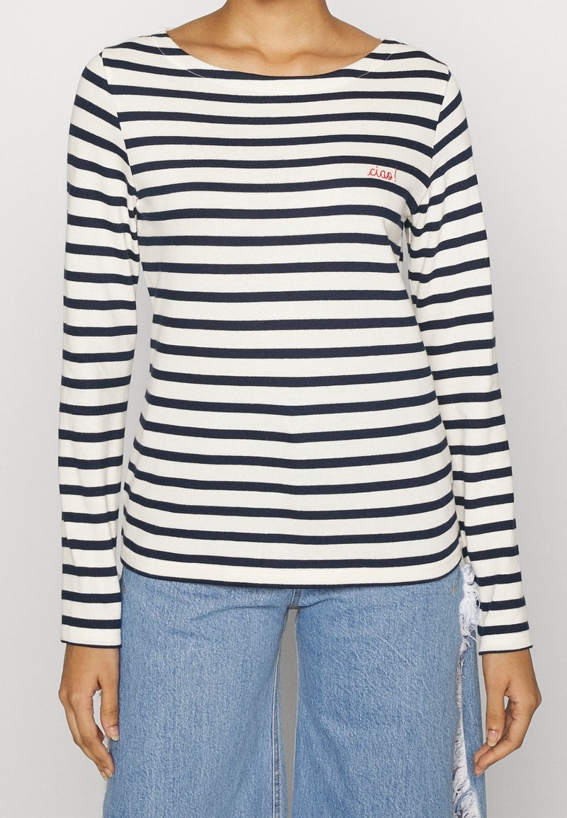 Osoba ubrana w biało-czarną koszulę w paski z długim rękawem, na której haftowane jest słowo "ciao!" w kolorze czerwonym oraz jasno-niebieskie, podarte jeansy.