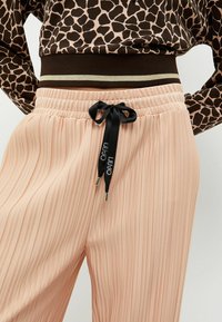 Pantaloni plissettati rosa chiaro con una cintura elastica, caratterizzati da un accento a righe marrone scuro e oro, e un cordino nero.