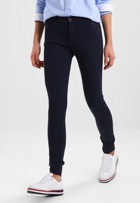 Marinblå stretchleggings med en åtsittande design, utrustade med en framficka med dragkedja och en dekorativ knapp. Kombinerat med vita sneakers.