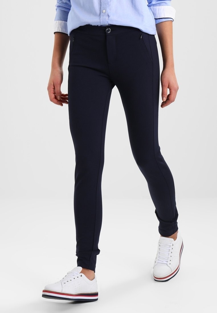 Marinblå stretchleggings med en åtsittande design, utrustade med en framficka med dragkedja och en dekorativ knapp. Kombinerat med vita sneakers.