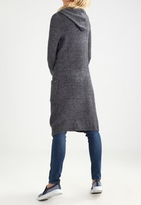 Długi szary kardigan z kapturem o miękkiej fakturze, wyposażony w boczne kieszenie i luźny krój. Noszony na niebieskich dżinsach i wzorzystych butach.