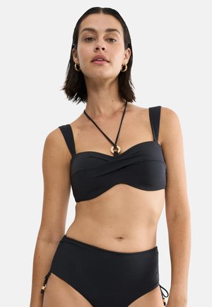 Kobieta w czarnym biustonoszu bikini typu bandeau z wiązaniem na szyi i wysokim stanem majtek bikini, na jednolitym jasnym tle.