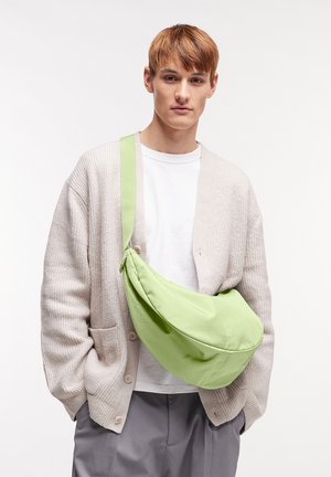 Groene stoffen schoudertas met een gebogen vorm, verstelbare schouderband en zachte textuur. Draagt over een wit shirt en een beige cardigan.