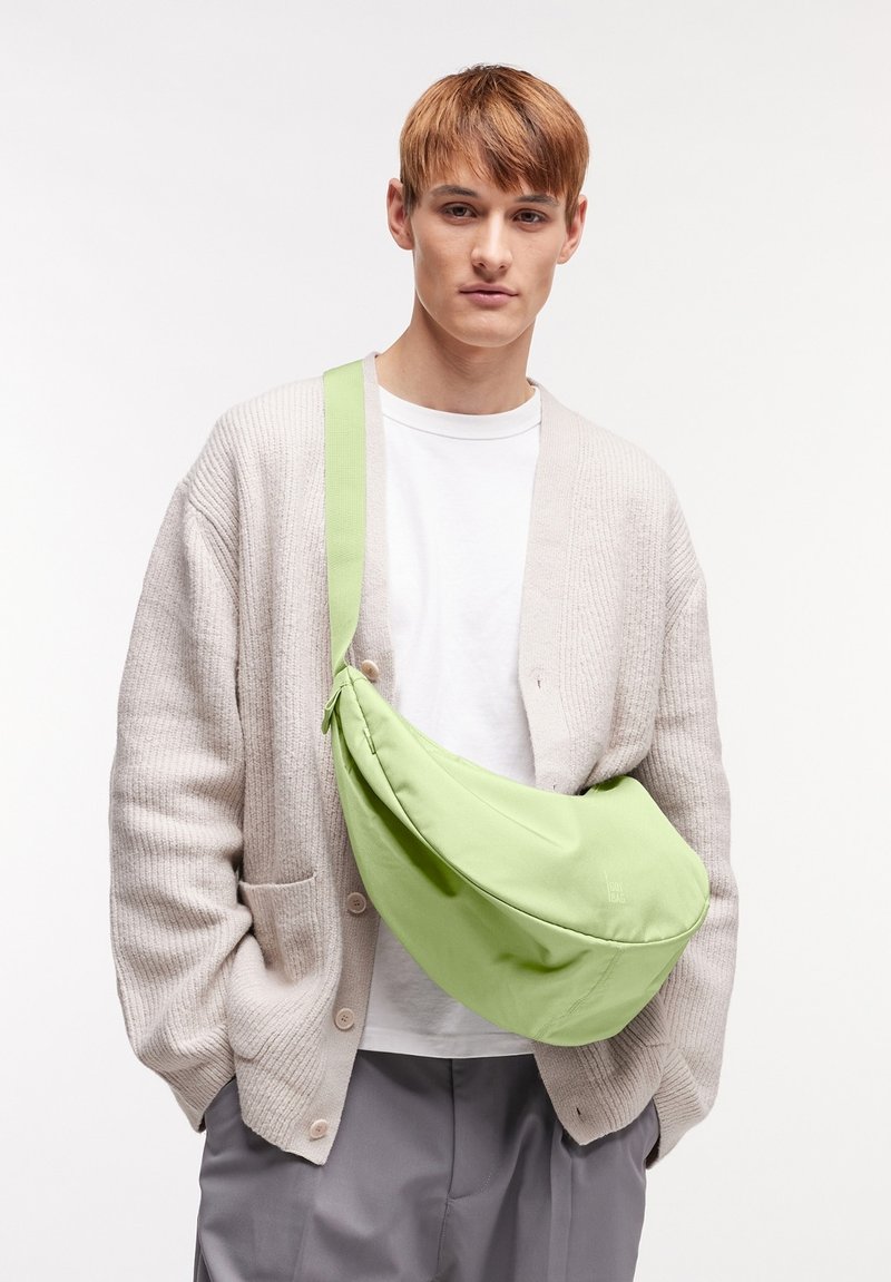 Borsa a tracolla verde in tessuto, con forma curvata, tracolla regolabile e texture morbida. Indossata sopra una camicia bianca e un cardigan beige.