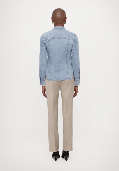 Lys blå denimjakke med en tettsittende design, bak sømmer og knapper på mansjettene, paret med beigh skreddersydde bukser og svarte hæler.