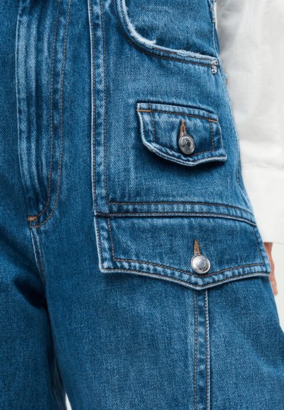 Κοντινή λήψη μπλε τζιν παντελονιού με διπλές τσεπές με καπάκι, ασφαλισμένες με ασημένια κουμπιά και εμφανείς καφέ ραφές.