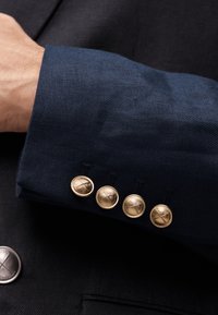 Nahaufnahme eines Marinejackenärmels mit vier goldenen Knöpfen, die gekreuzte Golfschläger zeigen, getragen über einem schwarzen Kleidungsstück mit einem einzelnen silbernen Knopf.