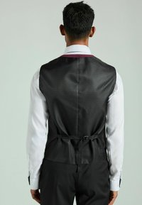 Chaleco negro perforado con un acento burdeos, que cuenta con una correa en la cintura. Combinado con una camisa blanca de manga larga y pantalones oscuros bien ajustados.