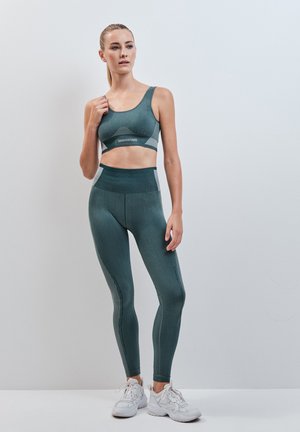 Donna che indossa un reggiseno sportivo verde scuro coordinato e leggings a vita alta, in piedi con scarpe da ginnastica bianche su uno sfondo chiaro e uniforme.