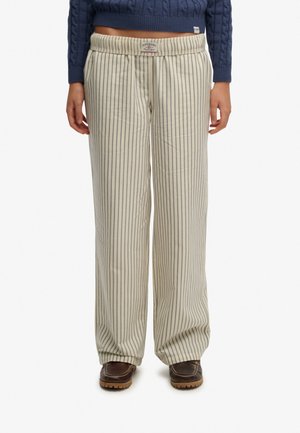 Osoba ubrana w beżowe spodnie o szerokich nogawkach z cienkimi pionowymi czarnymi paskami, brązowe buty i krótki granatowy sweter w warkocze.