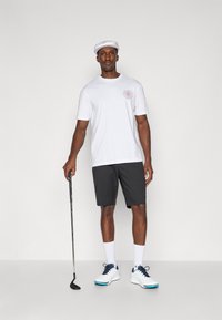 Camiseta branca de manga curta com um logotipo circular, combinada com calções pretos. O modelo usa sapatos de golfe brancos e um boné branco, segurando um taco de golfe.