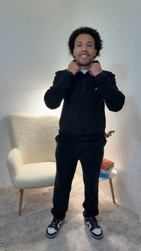 Black zip-up sweatshirt with a small logo on the chest, paired with black pants and black sneakers. The fabric appears soft and textured.