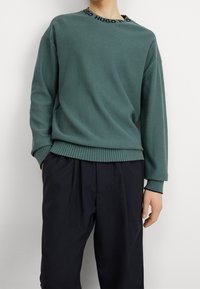 Homme portant un pull vert à manches longues avec "HUGO" sur le col et un pantalon noir ample, debout devant un fond blanc.
