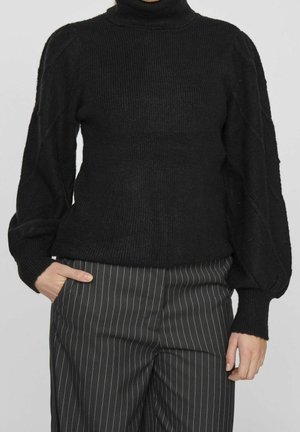 Persona con un suéter de cuello alto de punto grueso negro con mangas abullonadas y pantalones negros de raya diplomática, una mano en el bolsillo.