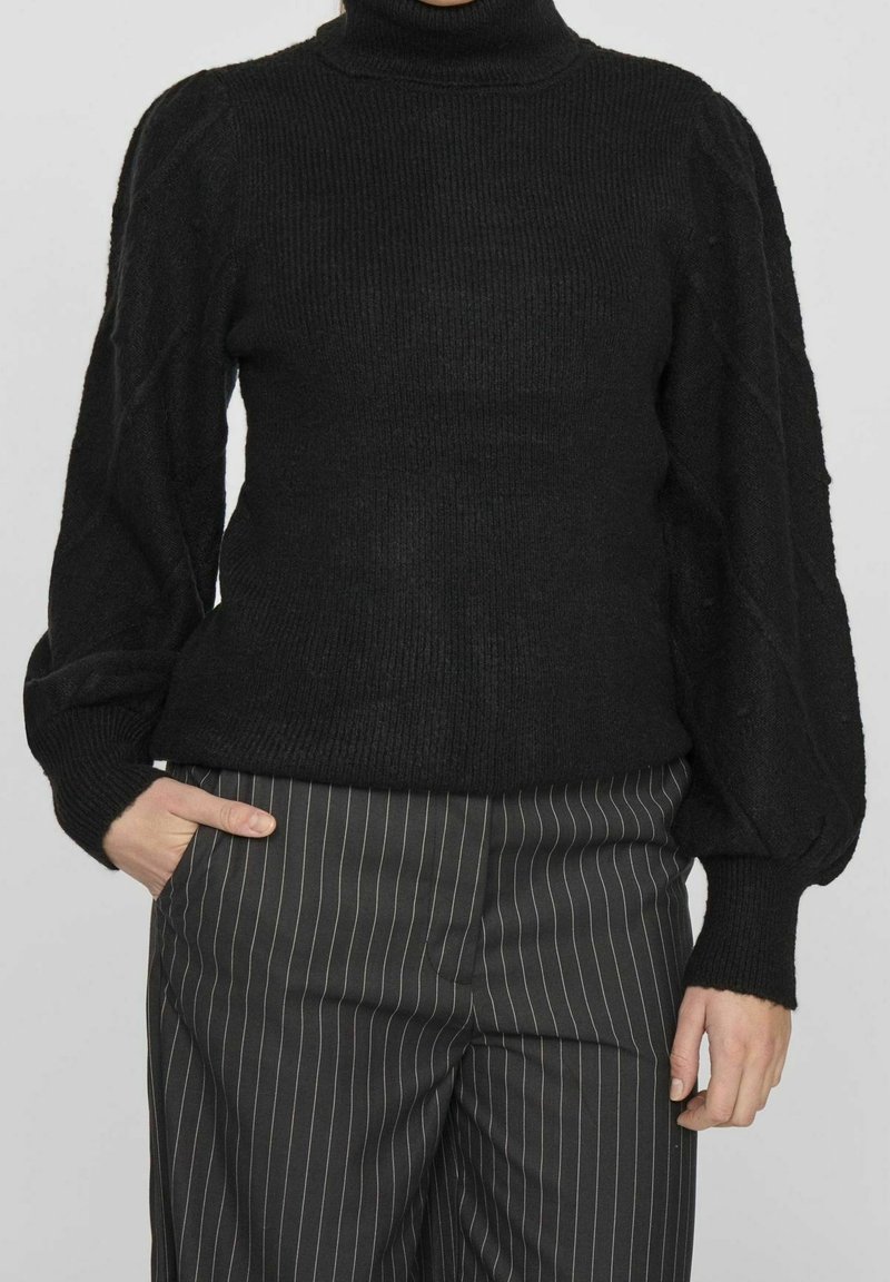 Personne portant un pull à col roulé noir en maille torsadée avec des manches bouffantes et un pantalon noir à fines rayures, une main dans la poche.