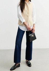 Beige ärmlös väst med knappar, vit skjorta under, mörkblå utsvängda jeans, svarta loafers och en liten svart handväska med ett guldspänne.