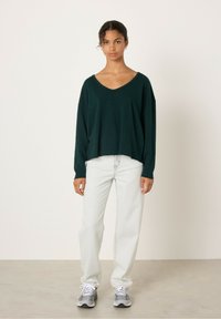 Tumši zaļš, pārmēru liels V veida kakla svīteris apvienots ar gaiši ziliem taisnā griezuma džinsiem un pelēkiem sporta apaviem, uz neitrālas fona.