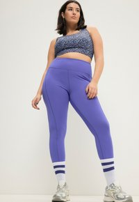 Paarse high-waisted leggings met stiksels, gecombineerd met een zwarte crop top met een blauw bloemmotief, en witte sportsokken met blauwe strepen.