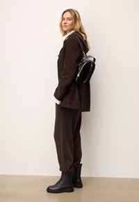 Hnedá semišová bunda, pod ňou biela košeľa s golierom, tmavohnedé jogger nohavice, čierne šnurovacie topánky, malá čierna taška so zlatými akcentmi.