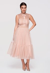 Femme portant une robe midi rose poudré sans manches avec des détails en dentelle et tulle, associée à des chaussures à talons hauts argentées et des boucles d’oreilles remarquables.