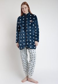 Chaqueta de forro polar azul marino con patrón de corazones blancos, cremallera frontal y bolsillo; combinada con pantalones de pijama gris claro que presentan un diseño de corazones similar.