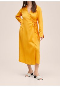 Gelbes Satin-Wickelkleid mit langen Ärmeln, verfügt über eine seitliche Bindung und einen vorderen Schlitz, kombiniert mit weißen Mules mit Blockabsatz.