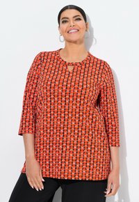 Nainen hymyilee, seisoo valkoista taustaa vasten, päällään korvakorut, oranssipohjainen kolmivartinen paita ja mustat housut.