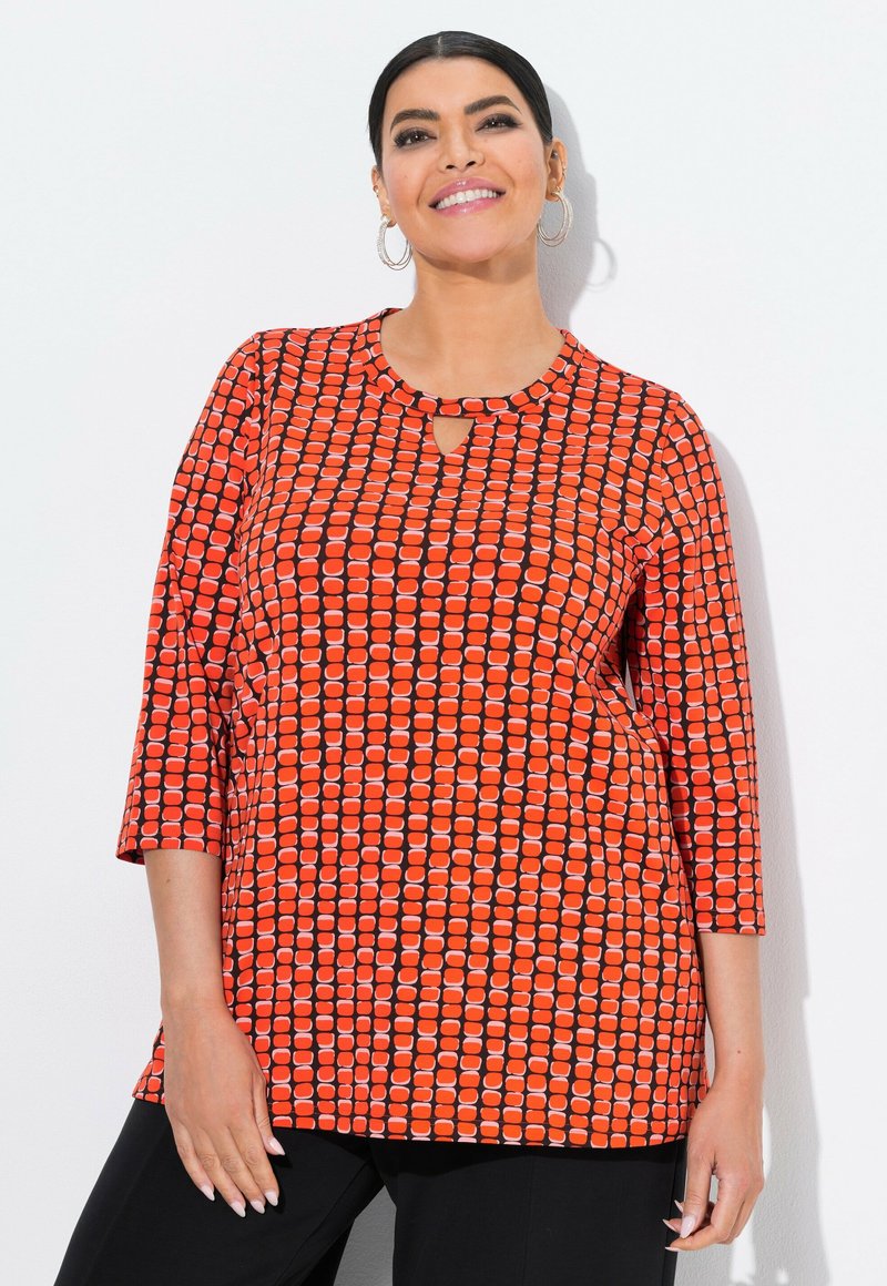 Nainen hymyilee, seisoo valkoista taustaa vasten, päällään korvakorut, oranssipohjainen kolmivartinen paita ja mustat housut.