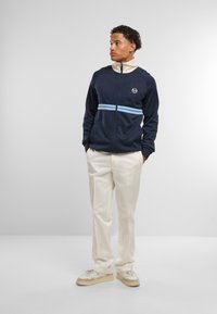 Mornarska zip-up jakna z svetlo modrimi črtami in logotipom, smetana visoki ovratnik, nosi se z smetanastimi hlačami in bež športnimi copati. Gladka tekstura tkanine.